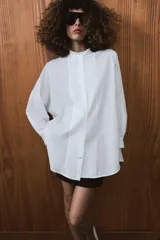 Camisa amplia de algodón con cuello redondo, manga por debajo del codo con puño y bajo asimétrico con aberturas laterales. Presenta cierre frontal con botones ocultos por solapa.