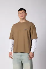 Remera de manga larga con diseño de doble capa, que simula una remera de manga corta color moka sobre una de manga larga blanca. Presenta un pequeño logo gráfico en el pecho y un detalle de etiqueta en el puño.