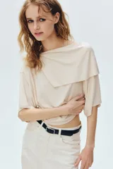 Top corto confeccionado en punto elástico de viscosa, color beige, con escote bote, hombros caídos y mangas cortas amplias. Presenta una sección superior superpuesta con un diseño asimétrico que crea un efecto capa.