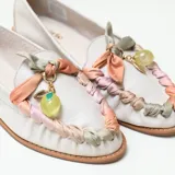 Mocasín de cuero color off-white vainilla, con taco bajo, punta redondeada y detalles artesanales de tiras entrelazadas en tonos pastel y aplique colgante con cuenta translúcida en el empeine.