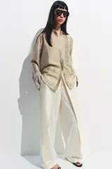 Camisa de cuello mao color beige, confeccionada en tejido satinado con brillo sutil, manga larga con puños anchos y un bolsillo de parche en el pecho con detalles bordados tonales. Cierre frontal con botones ocultos bajo solapa.