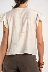 Blusa blanca sin mangas con escote en V, cierre frontal con botones y mangas con volados. Presenta pequeños bordados sutiles distribuidos por toda la prenda.