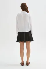Blusa blanca de viscosa con cuello a la base, mangas largas abullonadas con puños ajustados, detalles de volados, encaje y botones frontales.