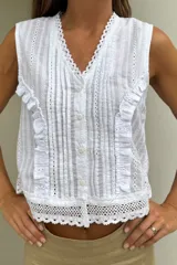 Blusa blanca sin mangas, de corte recto, con cuello en V, cierre frontal con botones y detalles de volados frontales y bordados calados.
