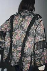 Kimono de tejido jacquard con estampado floral multicolor sobre fondo oscuro. Presenta un diseño abierto con aplicaciones de flecos negros a contraste en los bordes frontales y en las mangas. Posee mangas tres cuartos con terminación en vuelta y aberturas laterales en el bajo.
