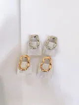 Par de aros colgantes con diseño geométrico compuesto por una pieza superior metálica de forma irregular y una pieza inferior cuadrada de resina con efecto nacarado.