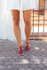 Sandalias planas estilo romano tipo cangrejera, confeccionadas en cuero charolado de color bordó. Presentan diseño de tiras entrelazadas en el empeine, puntera cuadrada, talón cerrado con tira ajustable mediante hebilla metálica dorada y suela plana con pequeño tacón.