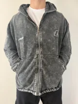 Campera con capucha y cierre frontal, de color gris con efecto lavado ácido. Presenta bolsillos frontales tipo canguro y logotipos bordados en el pecho.