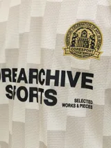 Camiseta tipo polo de manga corta con calce suelto. Presenta un diseño de cuadros en tonos beige y crema, con cuello y bordes de mangas en color negro. Incluye un escudo circular bordado en el pecho y estampado tipográfico en el frente.