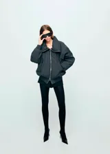 Chaqueta estilo bomber color negro, de corte oversize, con cuello alto amplio, cierre frontal de cremallera metálica y bolsillos laterales.
