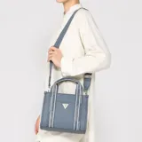 Cartera tipo tote bag Guess modelo Jackie de material sintético texturizado color celeste, con asas de mano y correa ajustable para el hombro con inscripciones de la marca. Cuenta con bolsillo frontal y aplique metálico triangular con el logo de Guess.
