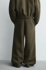 Pantalón de corte wide fit confeccionado en tejido técnico compacto con efecto neopreno. Presenta cintura elástica con cordón ajustable, pliegues frontales, bolsillos laterales y detalle de pliegues en las rodillas con aplicaciones metálicas.