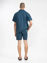 Bermuda de hombre color azul petróleo, confeccionada en mezcla de lino y algodón. Presenta cintura elástica con cordón, bolsillos laterales y trasero tipo parche. Incluye bordado floral en la pierna izquierda y etiqueta con logo en el bolsillo trasero.