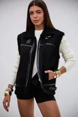 Chaleco negro de estilo aviador con interior y bordes de corderito sintético. Presenta cierre frontal metálico, detalles de tiras de cuero sintético y hebillas ajustables en el cuello y el ruedo.