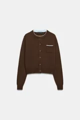 Chaqueta de punto color marrón con cuello redondo, manga larga y cierre frontal con botones. Presenta un bolsillo delantero tipo plastrón y detalles de ribetes en color celeste en el cuello y el bolsillo.