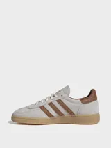 Zapatillas Adidas Handball Spezial de gamuza en tono gris claro, con las icónicas tres tiras laterales y refuerzo en el talón con textura de piel de serpiente en color marrón. Presenta suela de goma color caramelo y detalles dorados en el logo lateral y la lengüeta.