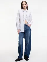 Camisa blanca a rayas verticales finas, con cuello clásico y cierre frontal con botones. Presenta apliques de pedrería en el cuello y en la tapeta de los botones.