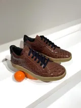 Sneakers acordonadas de cuero vacuno color marrón con textura de cocodrilo, suela plana color caramelo y detalles en negro.