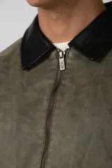 Chaqueta de estilo militar color verde oliva, confeccionada en gabardina resistente. Presenta cuello camisero en contraste color negro, cierre frontal con cremallera, dos bolsillos frontales tipo cargo con solapa y ajuste relajado.