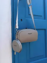 Bandolera compacta de cuero sintético con textura granulada, color beige. Incluye una correa ancha ajustable con un monedero circular desmontable y cierre superior con cremallera.