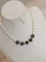Collar corto de cuentas cilíndricas blancas intercaladas con pequeñas esferas doradas y un grupo central de cuentas esféricas oscuras con vetas.