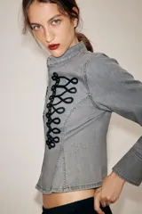 Chaqueta de denim gris con cuello subido y manga larga. Presenta un diseño entallado con bajo en evasé y un llamativo bordado frontal de alamares en color negro. Cierre frontal con corchetes ocultos.
