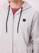 Campera canguro de felpa color gris claro melange, con capucha ajustable mediante cordones negros, cierre frontal completo, bolsillos tipo canguro y parche con logo de la marca en el pecho.