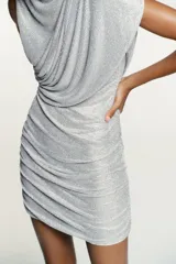 Vestido corto de tejido mesh plateado con aplicación de strass. Presenta cuello redondo, diseño sin mangas con drapeado en los hombros y una superposición efecto capa. Incluye frunces en los laterales y cierre oculto en el cuello.