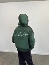 Buzo con capucha y cierre frontal completo, color verde oscuro con efecto lavado degradado. Presenta estampado gráfico en tipografía estilizada en el frente y mangas, bolsillos laterales y etiqueta de marca en el puño.