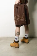 Bermuda estilo baggy de algodón color marrón oscuro, con cintura elástica y cordón blanco. Presenta un gran logo estampado en tono gris claro en la pierna derecha.