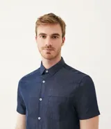 Camisa azul de manga corta con cuello clásico, corte slim fit, cierre frontal con botones blancos y bolsillo de parche en el pecho.