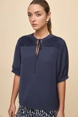 Blusa de manga corta color negro, con calce relajado, escote redondo con abertura y lazo para anudar. Presenta detalles de tablas horizontales en la parte superior que aportan textura al diseño.