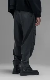 Pantalón de estilo utilitario confeccionado en textil técnico ripstop color gris oscuro. Presenta un corte oversize con cintura elástica, cierre frontal con broche técnico, bolsillos delanteros de gran tamaño con cierre y sistema de ajuste en los tobillos mediante broches snap.