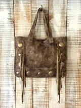 Bolso estilo shopper de cuero y gamuza en tono marrón desgastado. Presenta un diseño artesanal con flecos laterales largos y tachas metálicas circulares decorativas en los laterales y la base. Cuenta con doble asa superior para llevar al hombro.