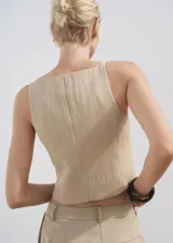 Chaleco tipo crop top de color beige, con tirantes finos, escote recto y cierre frontal de botonadura con botones revestidos. Cuenta con dos bolsillos de ribete delanteros y bajo con diseño asimétrico.