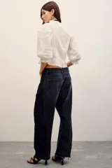 Pantalón de denim negro estilo barrel, de tiro medio con presillas y cierre frontal con botón y cremallera. Presenta un diseño de paneles en contraste a los lados y un corte amplio en las piernas con dobladillo deshilachado.