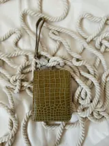Billetera pequeña de cuero con textura de reptil, color verde pistacho. Cuenta con cierre metálico perimetral, correa de muñeca extraíble y detalle de moneda metálica plateada con logo grabado.