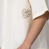 Camisa de manga corta color marfil, confeccionada en 100% algodón con calce boxy, cuello camisero, cierre frontal de botones y diseño con bordado sutil en la parte inferior.