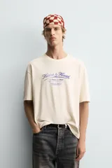 Camiseta de corte boxy fit, color beige con efecto lavado, cuello redondo y manga corta. Presenta un estampado gráfico en azul oscuro en el frente con tipografía estilo vintage.