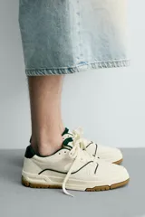 Zapatilla deportiva de estilo retro con diseño de paneles en tonos crema y blanco, detalles en verde oscuro y suela de goma color caramelo.