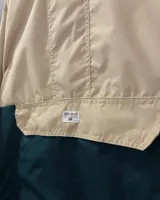 Campera rompeviento de diseño bicolor, con parte superior en tono arena y parte inferior en verde oscuro. Cuenta con capucha, cuello alto, cierre frontal parcial oculto y un amplio bolsillo frontal tipo canguro.