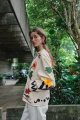 Chaqueta de algodón color crudo con cuello redondo, manga larga y bolsillos delanteros de plastrón. Presenta un diseño con bordados coloridos de flores y motivos abstractos a contraste, y cierre frontal con botones oscuros.