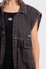 Chaleco de denim negro con efecto deslavado, diseño sin mangas, bolsillos frontales con solapa, costuras en contraste y botones metálicos.