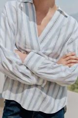 Blusa de manga larga confeccionada en mezcla de viscosa, lino y liocel, con diseño de rayas verticales en tonos celestes y marrones sobre fondo blanco. Presenta cuello solapa con escote en V cruzado.