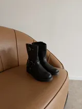 Bota de caña media en eco cuero negro, con diseño estilo biker que incluye correas con hebillas metálicas y herrajes plateados en el tobillo. Cuenta con suela plana y punta redondeada.