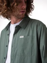 Camisa de manga larga convertible marca Caterpillar, color verde militar, con cuello camisero, cierre frontal de botones, bolsillo en el pecho con etiqueta de logo y dobladillo redondeado.