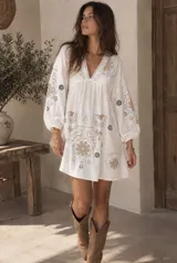 Vestido corto blanco de corte oversized, con escote en V y mangas 3/4 abullonadas. Presenta detalles bordados y estampados en tonos tierra y celeste en el frente y las mangas.