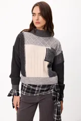 Sweater de punto con diseño patchwork en tonos gris, grafito y crudo, con cuello alto tejido. Presenta una estructura de bloques de color y texturas variadas, incluyendo detalles de trenzas y tejido acanalado.