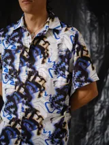 Camisa de manga corta de calce relajado, con cuello clásico, cierre frontal con botones y bolsillos aplicados en el pecho. Presenta un estampado integral abstracto en tonos crudo, negro, marrón y azul eléctrico.