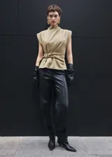 Blusa sin mangas color beige con cuello perkins y hombreras marcadas. Presenta un diseño recto con cinturón ajustable integrado en la cintura y cierre de cremallera en la parte posterior.